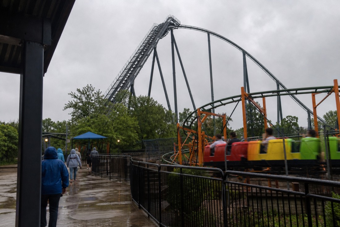 Rain-streaked coaster queue platform beside a tall idle roller coaster while a smaller ride keeps turning under heavy grey cloud